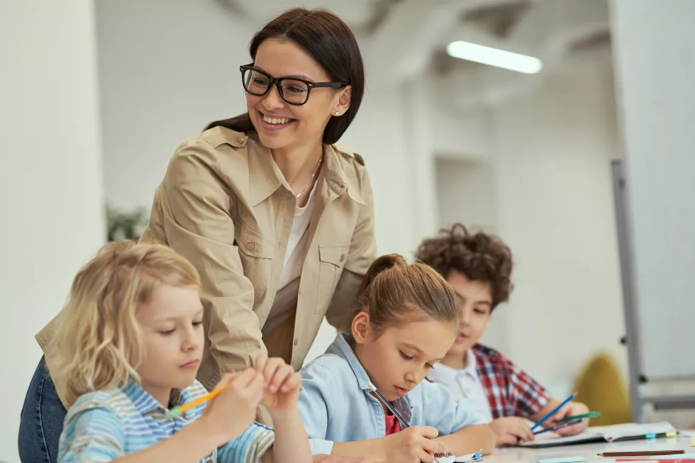 happy-young-female-teacher-glasses-helping-her-little-students-classroom-kids-sitting