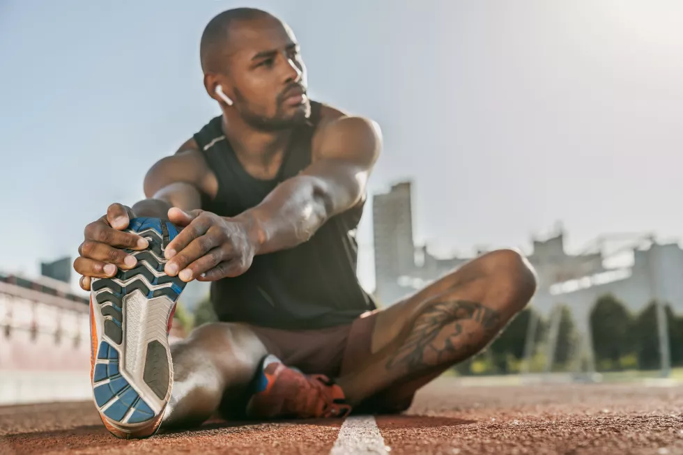 sporty-muscular-african-male-athlete-earphones-stretching-legs-while-sitting-stadium